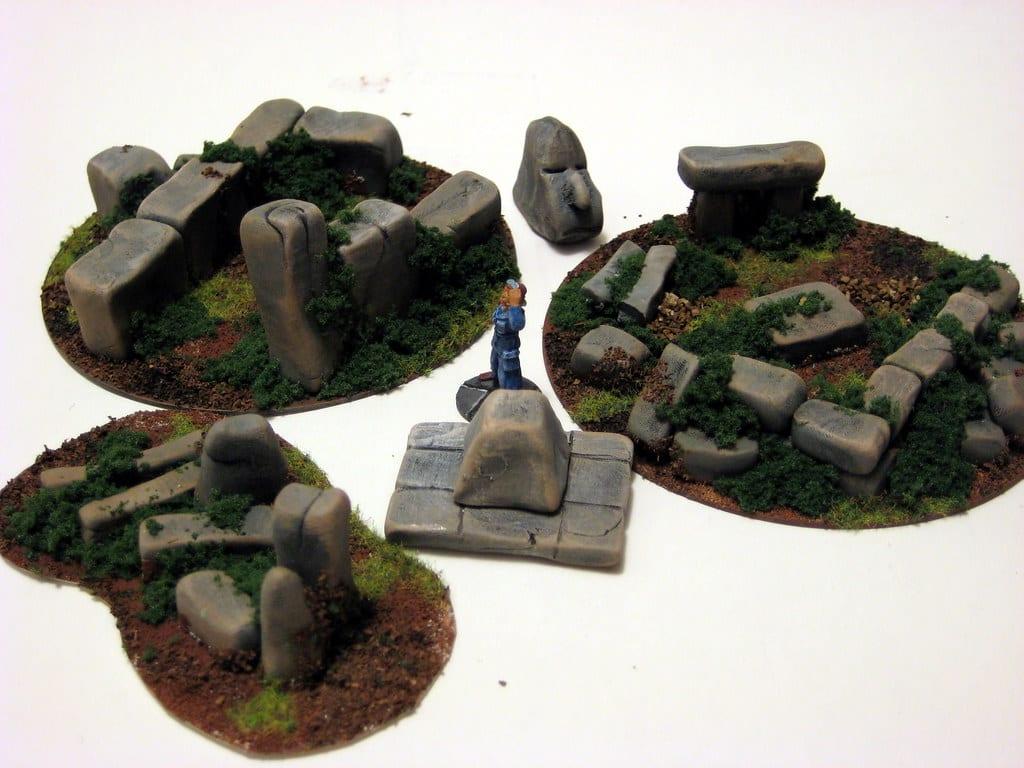 Outdoor scatter terrain pieces on flocked bases: painted standing stones, fallen boulders and a carved stone head, with a single miniature standing among them for scale