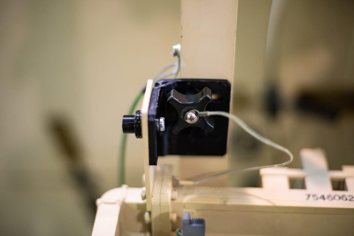 Black 3D printed bracket bolted onto an equipment frame, clamping a knob and cable in service