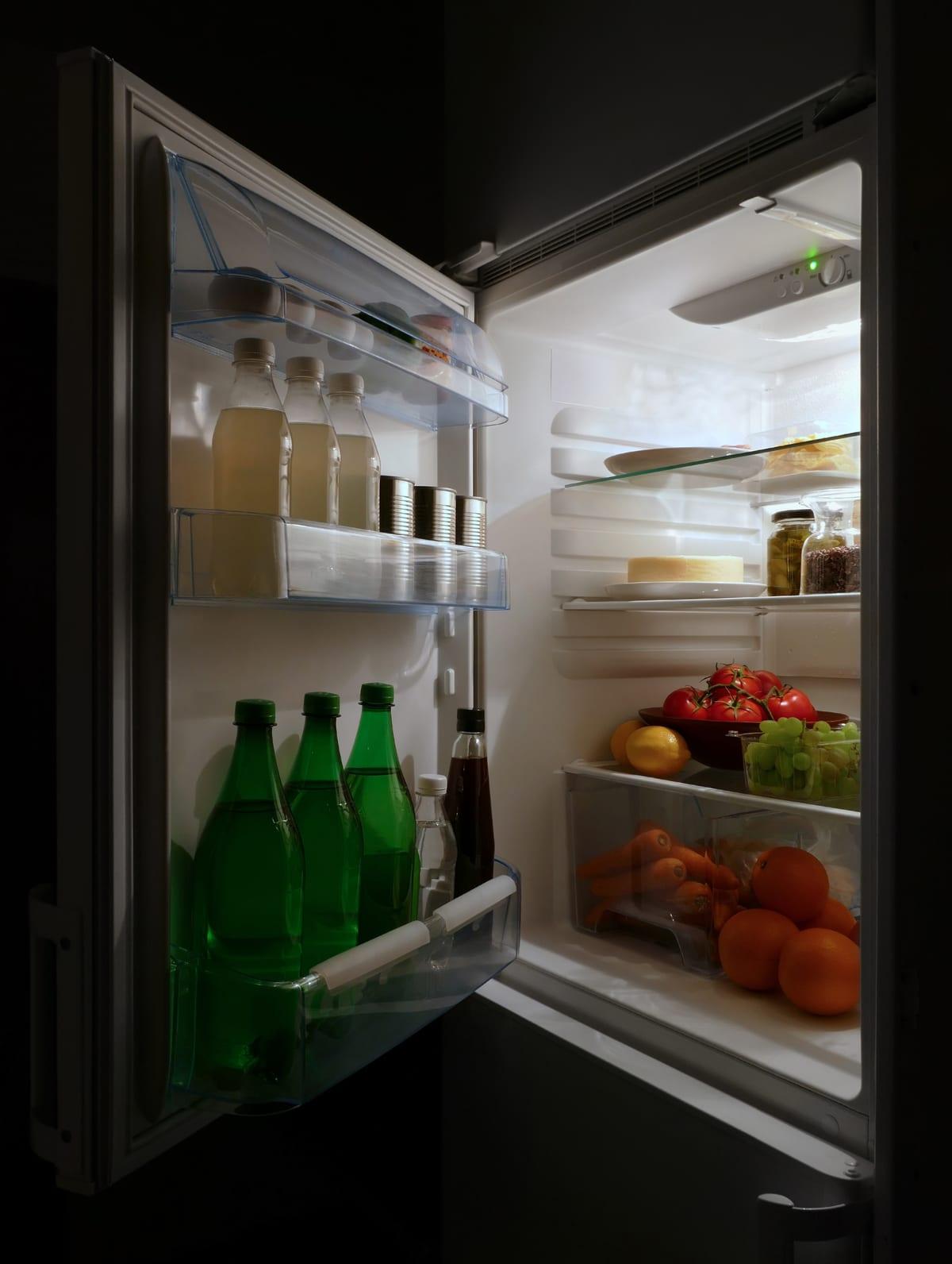 An open refrigerator at night with three door bins loaded with glass bottles and cans