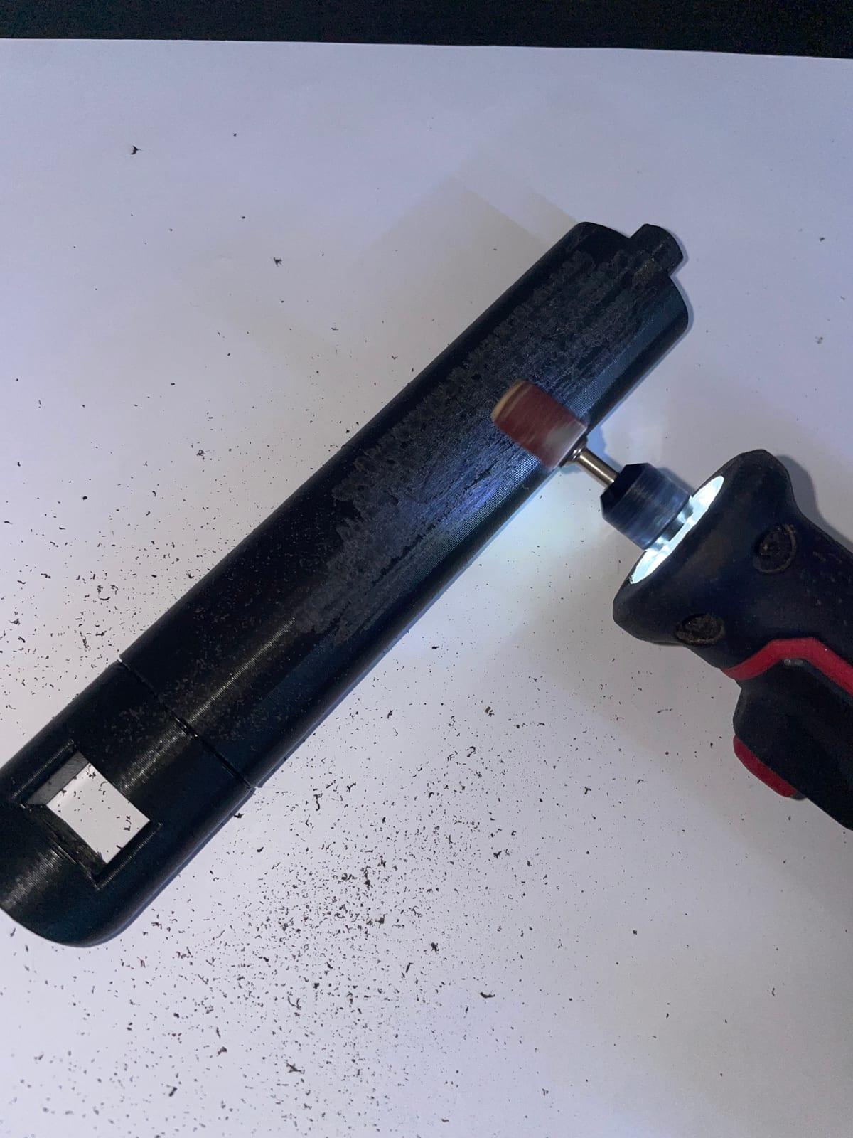 A black 3D printed part being smoothed with a rotary sanding tool, dust scattered on a white surface