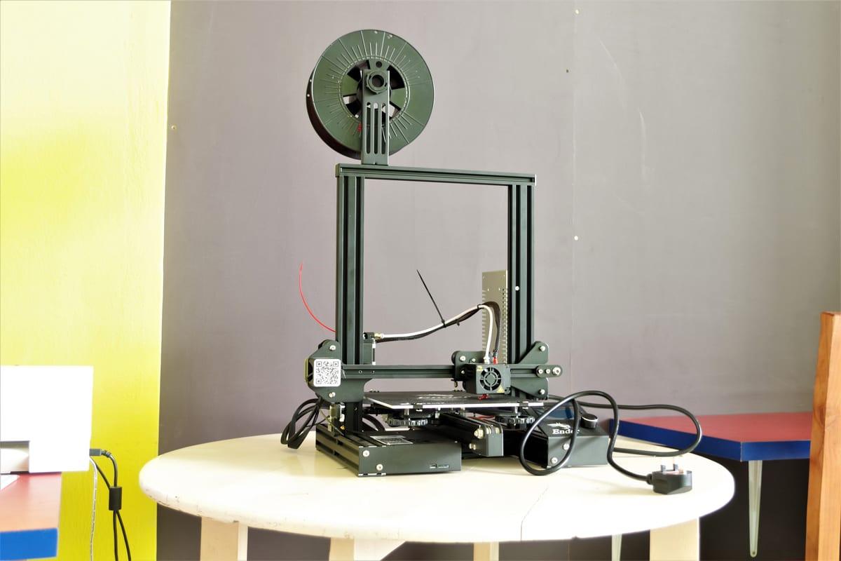 An open-frame desktop 3D printer on a small table with a spool of filament mounted above it