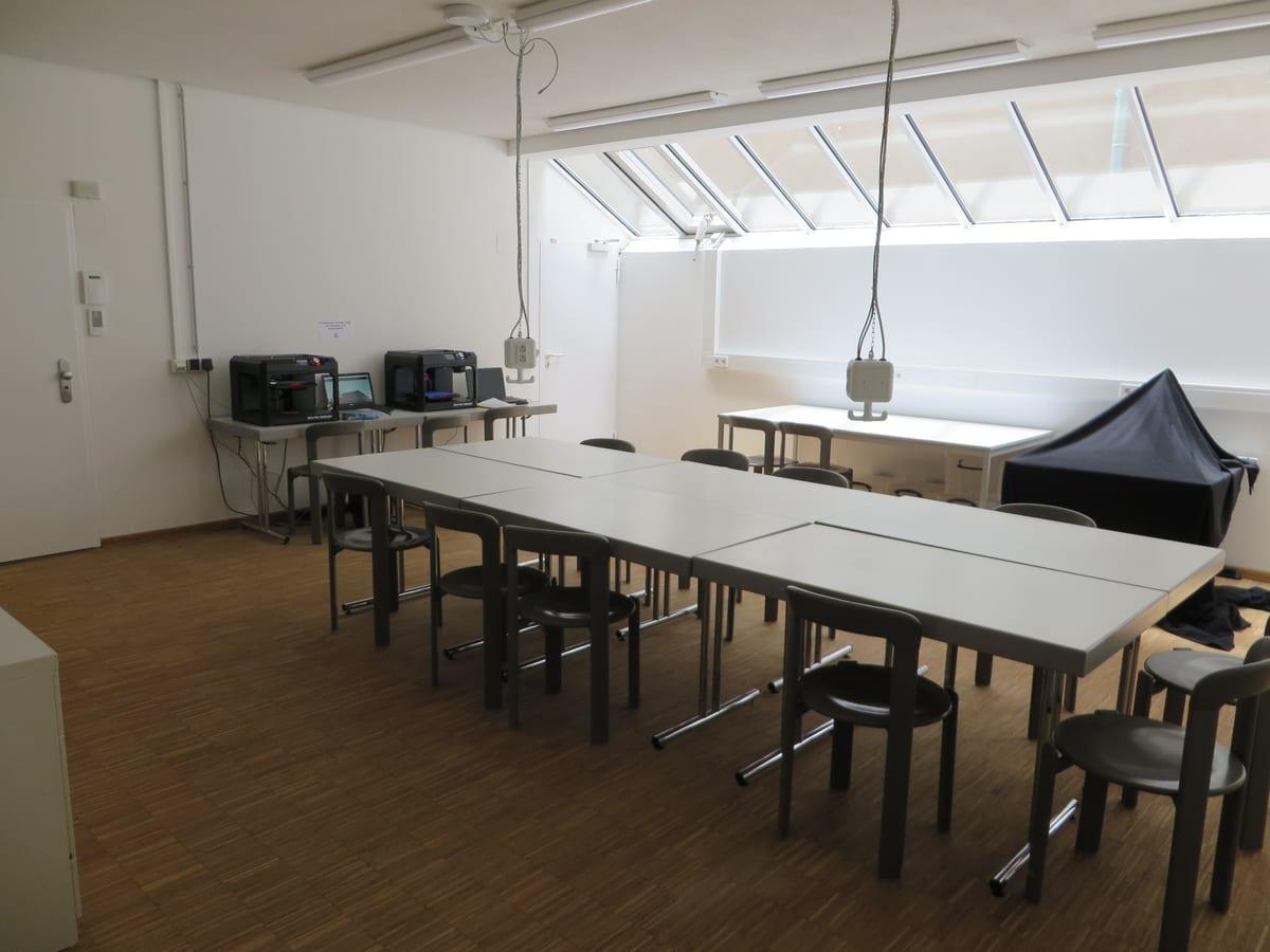 An empty public library makerspace room: two desktop 3D printers with laptops on a side bench, and grouped work tables with stools filling the rest of the room