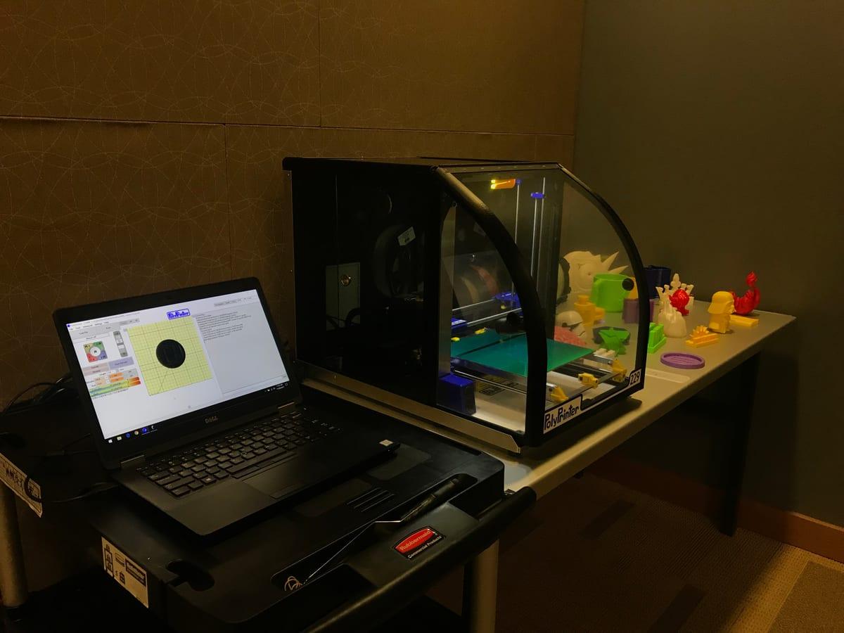 A laptop running slicing software beside an enclosed desktop 3D printer, with a row of printed models behind