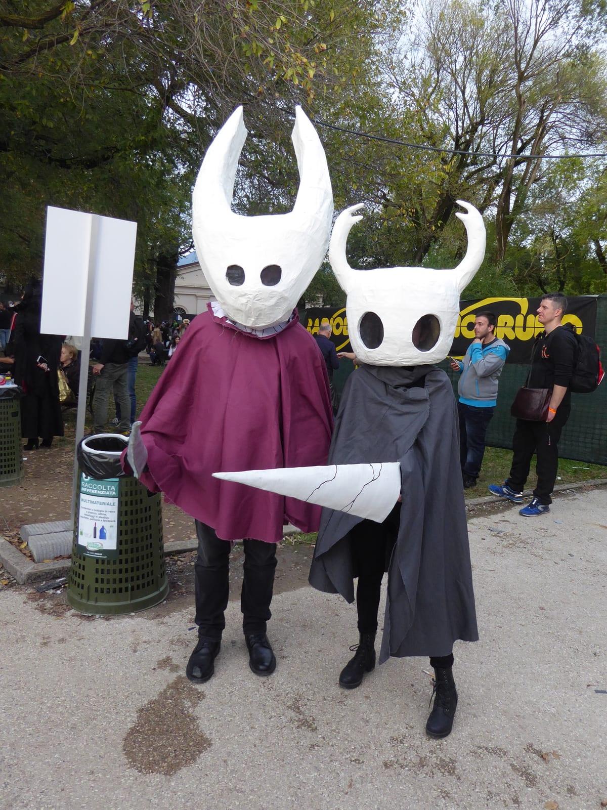 Two cosplayers wearing large smooth white horned character masks with cloaks
