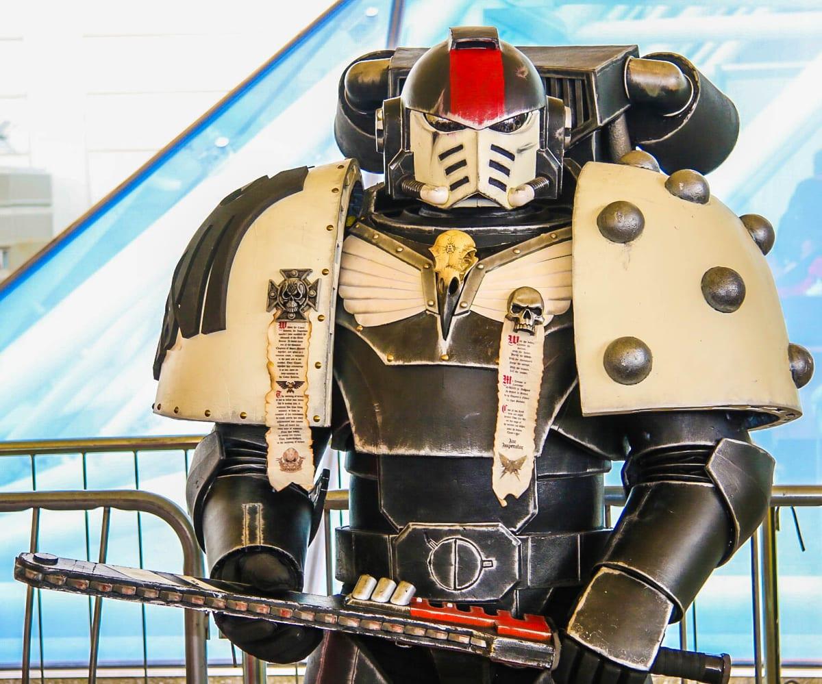 A cosplayer in oversized black and bone sci-fi power armour with huge rounded shoulder pauldrons, a helmet and a chainsword prop
