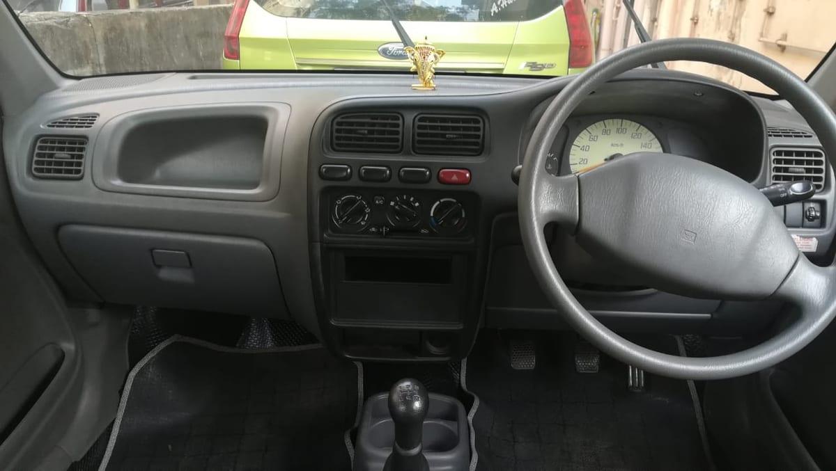 The grey moulded dashboard of a small right-hand-drive hatchback photographed from the back seat, showing the instrument binnacle, steering wheel, centre air vents, heater controls and gear lever