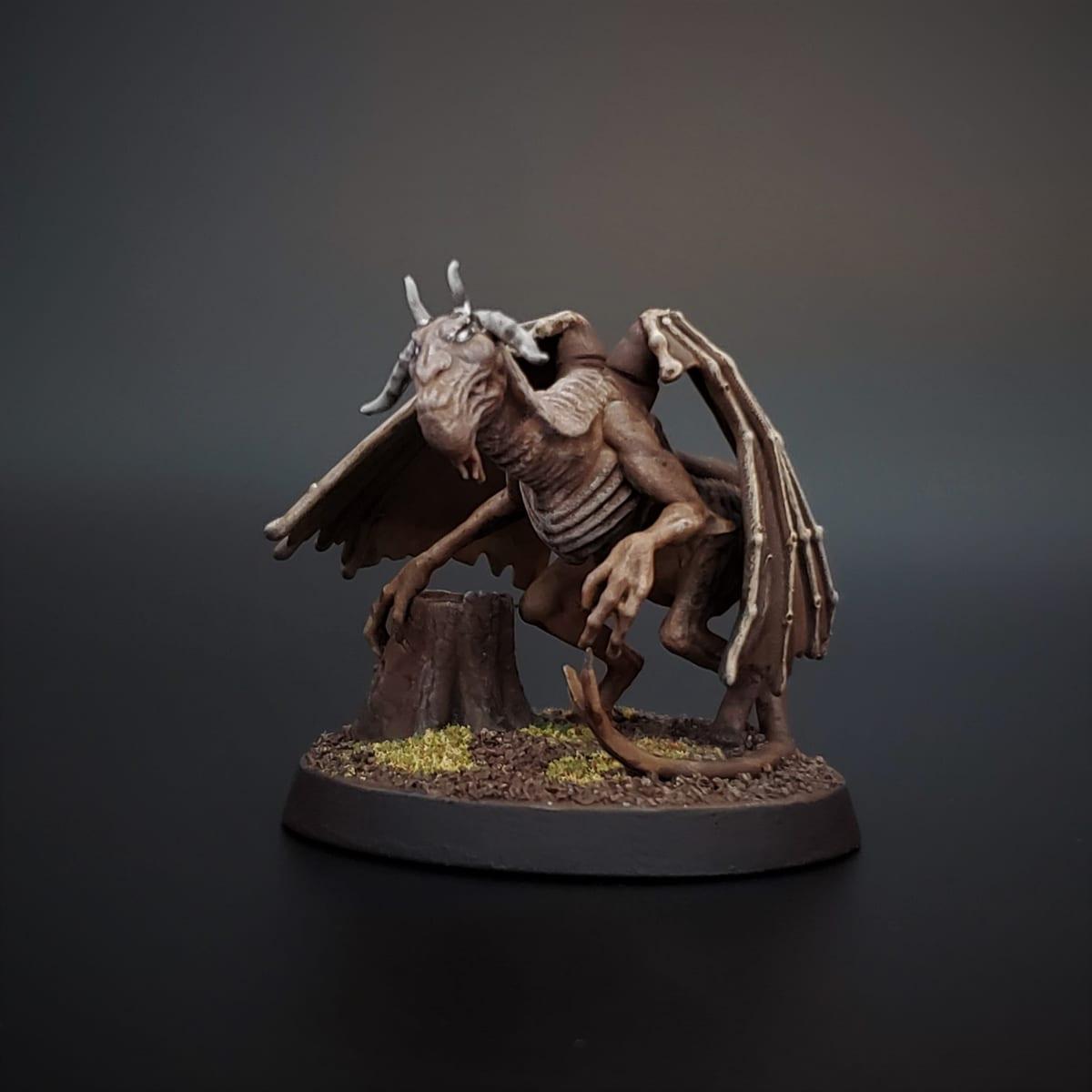A hand-painted tabletop miniature of a winged, horned creature perched by a tree stump on a round textured base, photographed against a dark background
