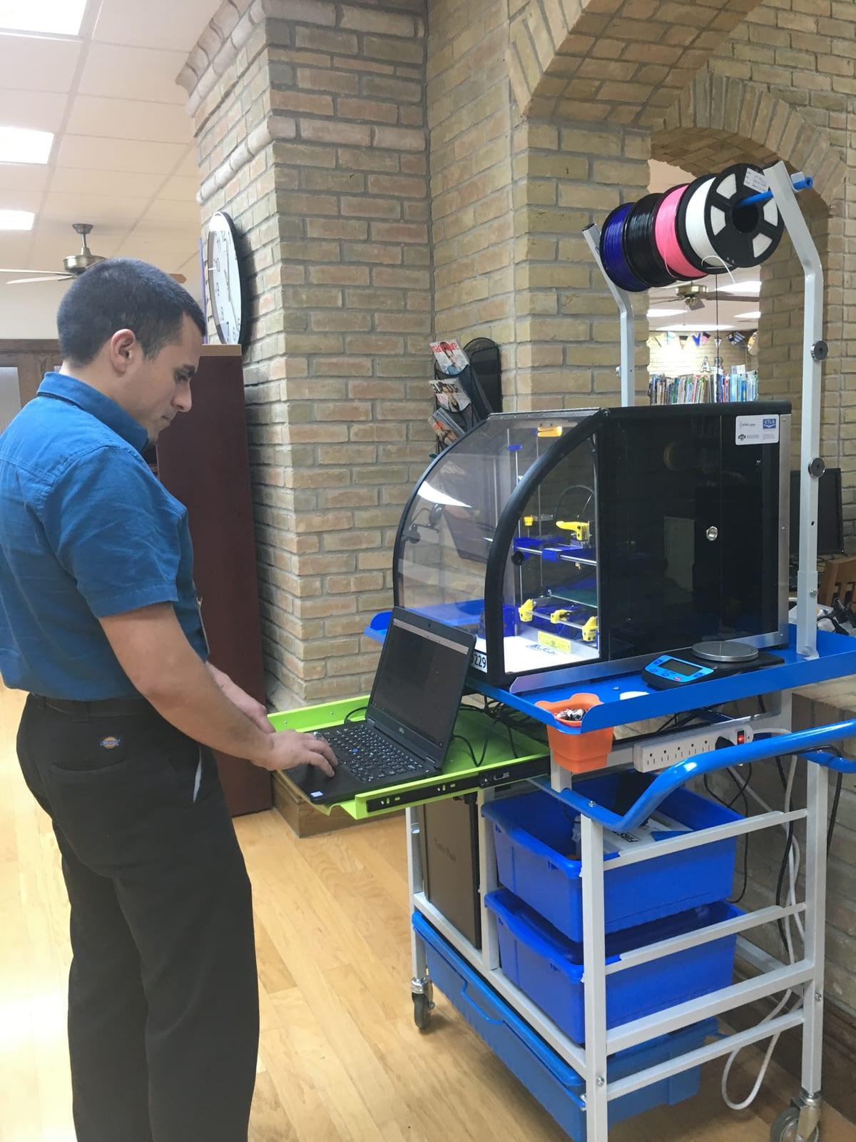 A library staff member in a blue shirt working at a laptop attached to an enclosed 3D printer on a wheeled cart, bookshelves and a wall clock behind him